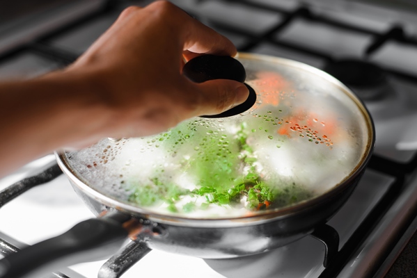 closeup of human hand opening frying pan with food on gas stove selective focus on misted glass lid of frying pan - Пицца из кабачков с колбасой