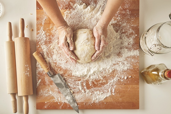 top view on white table with isolated wooden board with knife two rolling pins bottle olive oil transparent jar with flour woman hands hold prepared dough for pasta or dumplings - Пицца постная на тонком тесте