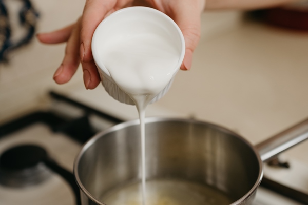 a photo of a hand of a woman pouring cream from the cup to the saucepan with lemon juice and zest on the gas stove - Драчена на сметане и сливках