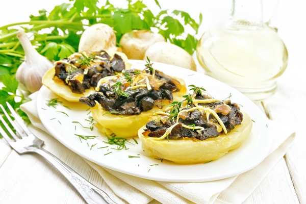potatoes stuffed with mushrooms fried onions and cheese in a dish on kitchen towel vegetable oil in decanter parsley garlic and a fork on wooden board background - Фаршированный картофель, постный стол