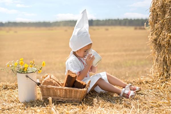 a girl in national belarusian clothing sits in a field on a stack of straw and drinks milk from a jar - Минский хлеб