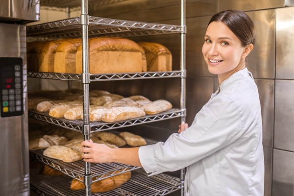 freshly baked bread pretty woman turning her face to camera standing near oven and trays of bread in great mood - Ржаной столовый хлеб