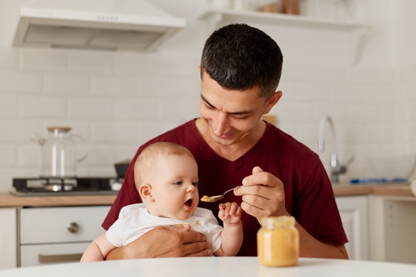 indoor shot of happy father sitting at table with little baby girl in arms and feeding daughter with fruit or vegetable puree complementary feeding of child - Средиземноморская диета