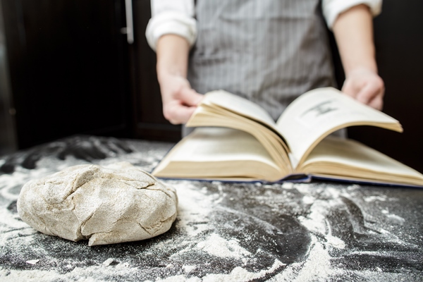 kneaded dough lies on table sprinkled with flour against of a baker leafing through a cookbook - Ржаной столовый хлеб