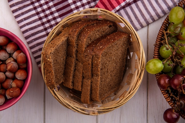 top view of rye bread slices in basket on plaid cloth with bowl of nuts and basket of grapes on wooden background - Хлеб бородинский штучный, высший сорт