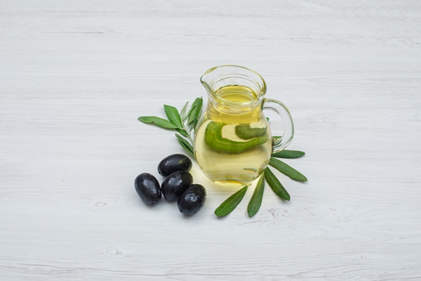 black olives and olive oil in a glass jar with olive leaves side view on white wood plank - Оливковое масло