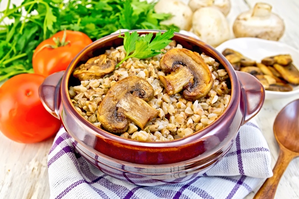 boiled buckwheat with mushrooms in a brown pottery bowl on a kitchen towel parsley tomatoes and fried mushrooms on the background light board - Углеводная питательность рациона