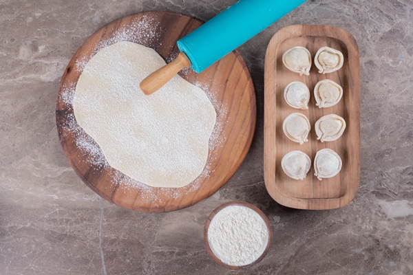 raw dumplings on wooden board with dough and flour on marble - Тесто на кефире для пельменей и вареников