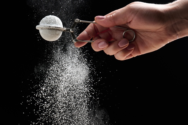 female hand holds a metal strainer with powdered sugar particles fly in different directions on a black background sprinkling - Постная галета с чёрной смородиной и яблоком