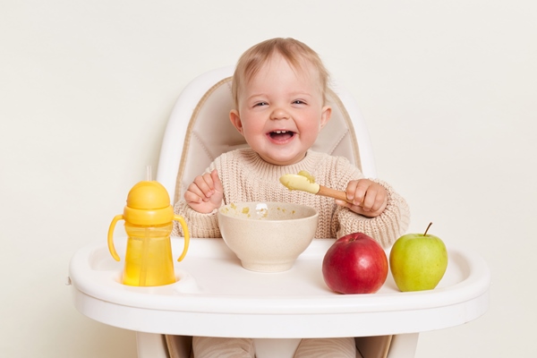 image of deligted laughing infant baby girl dresses in beige jumper sitting in high chair and eating isolated over white background holding spoon in hands enjoying tasty fruit puree - Организация правильного питания детей