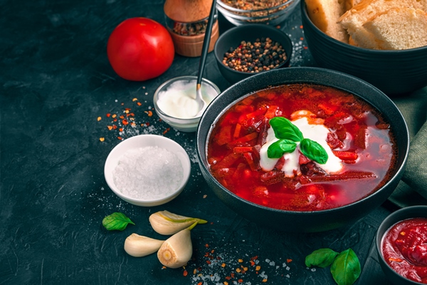 a black plate with fresh borscht on a background of ingredients on a concrete black background side view with copy space - Свекольник со сметаной (школьное питание)