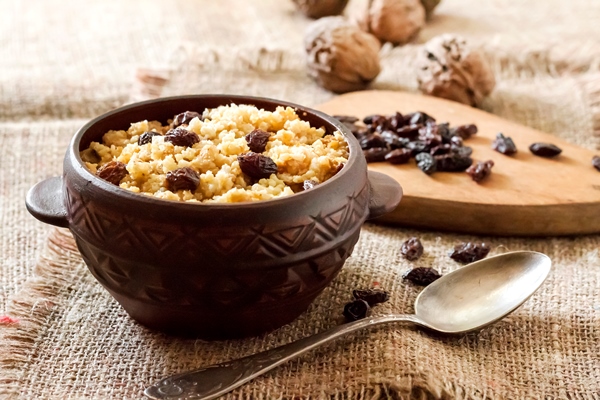 sweet millet porridge with dark raisins in ceramic rustic bowl with walnuts on background - Кукурузная молочная каша с изюмом, вязкая (школьное питание)