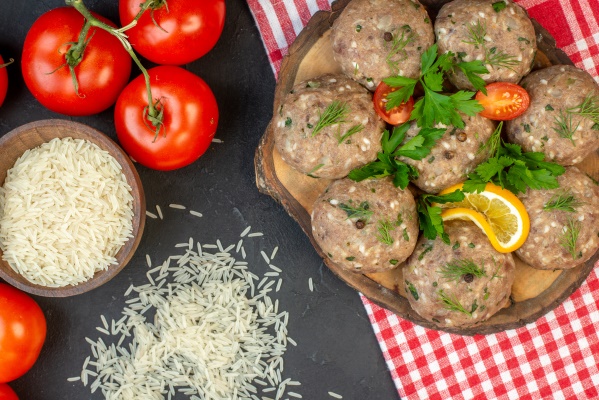above view of uncooked meatballs on a wooden cutting board rice inside and outside of a brown pot tomatoes with stems on dark wave background 1 - Тефтели с рисом "Ёжики" (школьное питание)