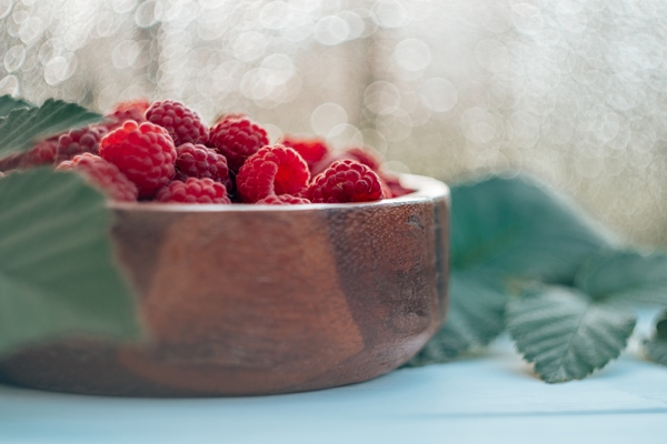 partially blurred wooden round bowl full of ripe red juicy raspberries on white wooden surface against bokeh background with raspberry bush leaves - Компот из малины (школьное питание)