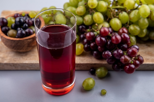 side view of grape juice in glass and grapes with bowl of grape berries on cutting board on gray background - Душепарка безалкогольная