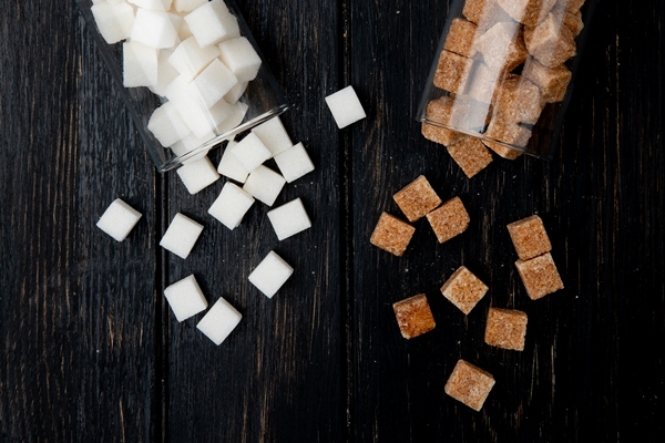 top view of white and brown sugar cubes scattered from glass jars on dark wooden background with copy space - Компот из смеси сухофруктов (школьное питание)